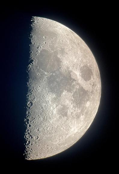 A first quarter moon against an almost black sky. Numerous craters are visible at the terminator, on the right, due to the low sun angle affording good shadow detail. Dark maria are prominent on most of the rest of the moon’s visible surface, against the lighter surrounding areas