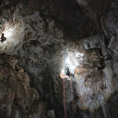 A person with a headlamp abseiling into a large cave at least 50m high. The persons shadows from two lamps are on the back wall with rusty-brown limestone and white comcretions