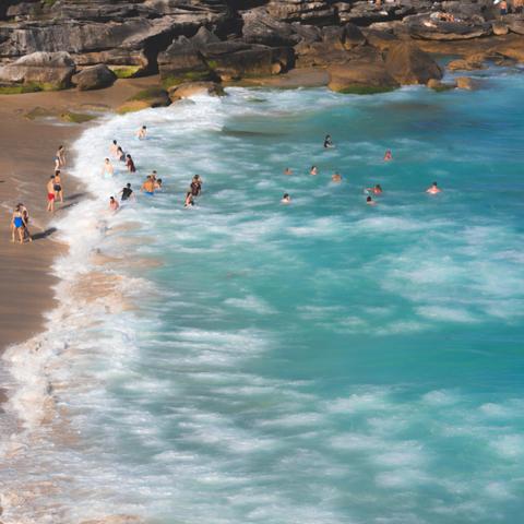 Waves gently wash over a sandy beach where several people wade and swim in clear turquoise water. Rocky formations line the upper edge of the beach. Some individuals stand near the shore, while others enjoy deeper waters. The scene is sunlit, indicating a clear day.