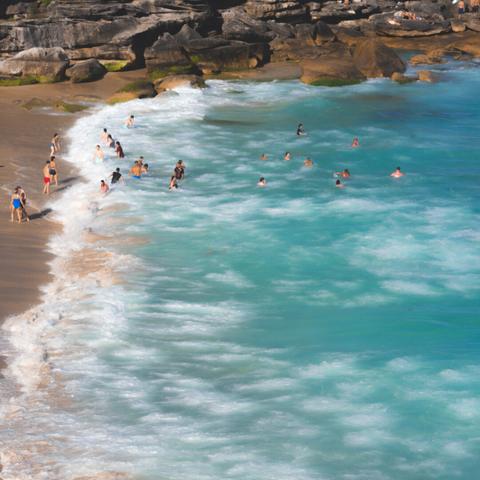 Waves gently wash over a sandy beach where several people wade and swim in clear turquoise water. Rocky formations line the upper edge of the beach. Some individuals stand near the shore, while others enjoy deeper waters. The scene is sunlit, indicating a clear day.