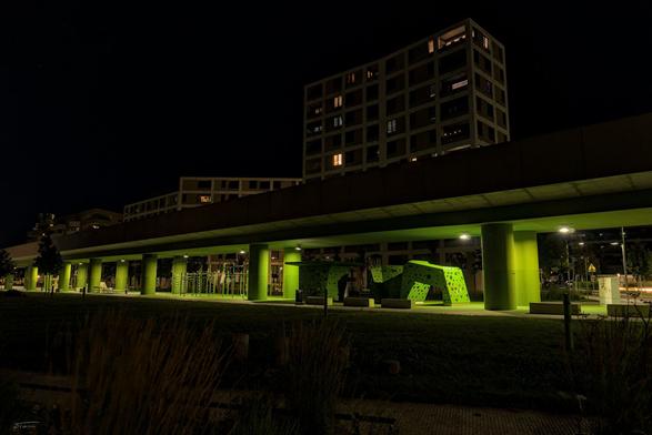 Auf dem Foto ist ein nächtlicher Blick auf einen Sport- und Kletterpark in der Seestadt Aspern (Wien) zu sehen, der sich unter einer erhöht verlaufenden U-Bahn-Trasse befindet.
Die Szene ist bei Nacht aufgenommen, was durch die dunkle Umgebung und die punktuelle Beleuchtung deutlich wird.
Die U-Bahntrasse verläuft waagrecht durch das Bild, gestützt von leuchtend grün angestrichenen Säulen, die den Park darunter strukturieren.
Unter der Trasse befinden sich verschiedene Sportgeräte, darunter ein Outdoor-Fitnessbereich (links) und mehrere grüne Kletterelemente mit bunten Griffen (rechts).
Im Hintergrund sind moderne Wohngebäude mit beleuchteten Fenstern zu erkennen, typisch für die neue urbane Architektur der Seestadt.
Das Licht der Laternen erzeugt scharfe Kontraste und hebt die grün gestrichenen Bereiche visuell hervor.