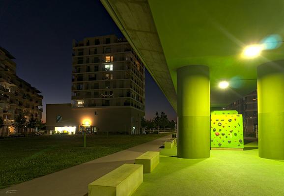 Dieses Bild zeigt dieselbe Szene aus einer anderen Perspektive, diesmal mehr von der Seite und mit einem stärkeren Fokus auf den Bereich direkt unter der U-Bahn-Trasse in der Seestadt Aspern bei Nacht.

Im Vordergrund dominieren die massiven, grün gestrichenen Säulen, die die Hochtrasse der U-Bahn tragen.
Direkt unter der Trasse sieht man einen grünen Kletterwürfel mit bunten Griffen, beleuchtet von mehreren starken Deckenlampen, was die Farben intensiv hervorhebt.
Links öffnet sich das Bild auf einen Grünbereich mit Rasen und einem Weg, der ins Wohngebiet hineinführt.
Am linken Bildrand sind moderne Wohnhäuser mit vielen Balkonen zu sehen, einige mit Lichterketten oder beleuchteten Fenstern.
Im Hintergrund erkennt man ein hell beleuchtetes Geschäft mit dem Schild "bolderbar", das eine warme Atmosphäre in die nächtliche Szene bringt.
Der Himmel ist bereits tiefblau, was auf die blaue Stunde oder den späten Abend hinweist – ein stimmungsvoller Übergang zwischen Tag und Nacht.
Diese Perspektive betont besonders die architektonische Wirkung der U-Bahntrasse und das Zusammenspiel von urbanem Raum, Licht und öffentlichem Sportangebot.