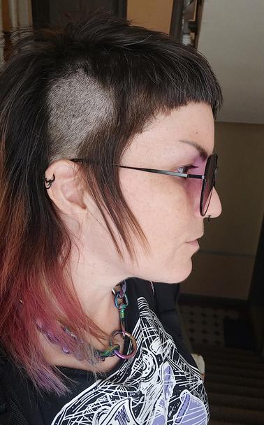Profile photo of a white non-binary person. Their haircut is a mullet (short at the top, long at the back), part of the side of the head is shaved. The ends of the long hair at the back have some leftover pink dye. They are wearing sunglasses.