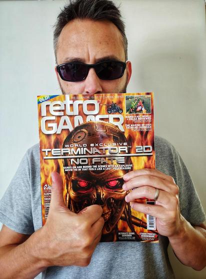 A white man wearing dark sunglasses and a grey t-shirt, holding a retro computer magazine (which features "Terminator 2D: No Fate", the upcoming game from the Bitmap Bureau team), while also giving a thumb-up. Wonder what the references could be... 🤔😉