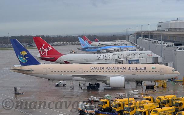 Side view of a twin engined jet airliner parked nose in to a grey terminal building.
The plane is mostly a sandy brown, with a thin, golden pinstripe separating that from the grey belly.
There are dark blue "Saudi Arabian" titles on the upper forward fuselage, in an Arabic script at the front, and in English towards the rear.
The tail is dark blue, with a thin, golden crescent moon shadowed in light blue, with a pair of crossed golden cutlasses under a golden palm tree.
Damp, grey, concrete apron fills the foreground, with a small fleet of yellow trucks in the bottom right corner of the frame.
A grey terminal building lines the right of the frame, with several other jet airliners, some white and red, others various shades of blue, white, and red, parked against it.
Trees run across the frame in the distance, under grey skies.