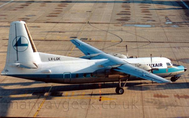 High side view of a high-winged, twin propellor-engined airliner facing to the right but being pushed back to the left by an out-of-frame tug attached to the nose-wheel by a long blue pole.
The plane is mostly white, with a grey belly, and a thick, light blue stripe running along the body.
There are black "LuxAir" titles on the upper forward fuselage, between the cockpit windows and the wing.
The tail is white, with the blue outline of a circle containing a rather angular, blue, stylised flying bird, and the black registration "LX-LGK" on a fillet at the front.
Grey concrete apron with occasional damp patches fill the rest of the frame.