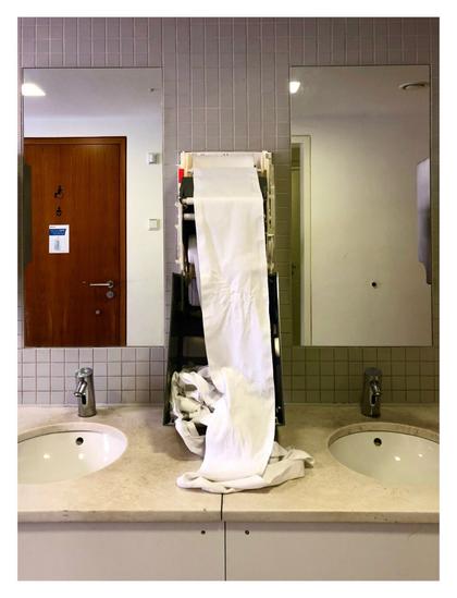 Foto im Hochformat. Waschbecken, Spiegel und Handtuchautomat in einer öffentlichen Toilette. Der Handtuchautomat ist geöffnet und die Papierhandtuchrolle hängt meterweit heraus. Der Automat ist genau mittig im Bild, links und rechts sind Spiegel in denen sich Türen spiegeln.