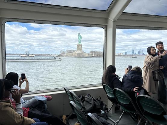 Cruise ship with angled windows, the Statue of Liberty in the background. People on the cruise are taking pictures, a selfie…