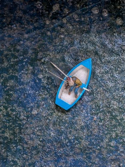 This is an overhead shot looking down into a body of water, where a small blue boat with two people is central to the image.
The water itself is a dark, murky blue with what appears to be a textured surface, perhaps from reflections, ripples, or even some form of debris or plant life on the surface, creating a somewhat mottled and uneven appearance. There are lighter patches within the darker blue, adding to the texture.

Märklin World Titisee.
