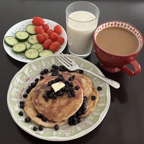A plate of buckwheat pancakes topped with a slice of butter, blueberries, and raspberry syrup. A side plate of slice cucumbers and whole grape tomatoes, and a cup of almond milk and almond coffee