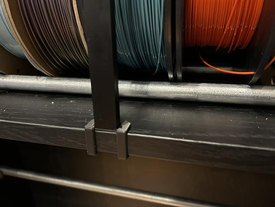 The black center shelf of an IKEA BILLY bookcase has a 3D printed plastic cap that wraps around its front edge.  Sitting in a niche in the clip is an aluminum C section aluminum channel, open side facing inward.  Behind are steel rails and several rolls of printer filament.