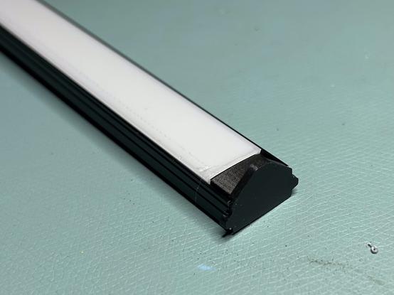 The aluminum channel is on a workbench. A 3D printed plug is inserted into the end.  Past the plug, the open side of the channel is covered by a milky transparent diffuser.  There is no provision for wires to get into the channel.