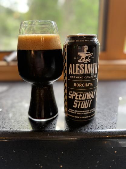 A stout glass full of dark beer with a thick tan head. The black can has white lettering with an image of an anvil and the AleSmith logo