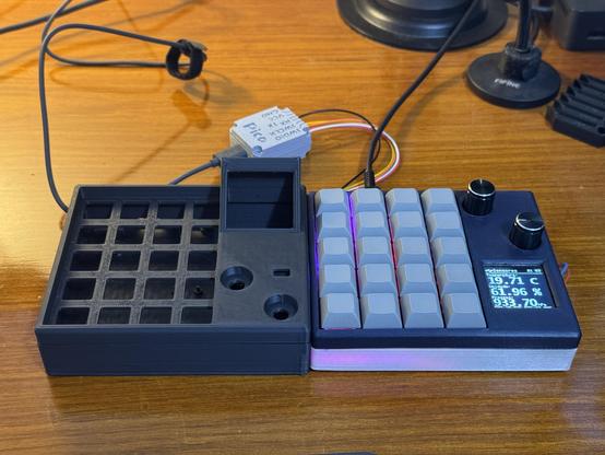 Two Macropad Pads. The one in the right is fully assembled, while the other in the left with a slightly tweaked design has only a 3D printed shell. They are both in a table, and there are wires plugging in the Macropad on the right