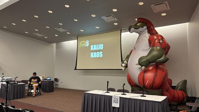 A photograph of a big anthropomorphic inflatable sockeye salmon next to a screen in a DLCC lecture room.  A man in a wheelchair is to the left of the image.