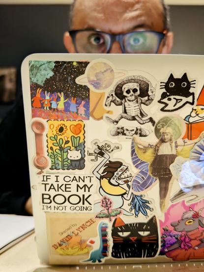 Sobre una mesa, la tapa abierta de una laptop muestra muchas calcomanías pegadas. Detrás, un poco desenfocada, se ve la cabeza de un hombre con lentes.