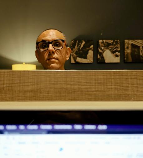 En el borde de un espejo, delante de una lámpara y una fila de fotografías fijadas a una pared, asoma la cabeza de un hombre con lentes y el cabello muy corto. Delante del espejo, muy desenfocada, se ve la pantalla de una laptop y, en ella, la ventana de una aplicación.