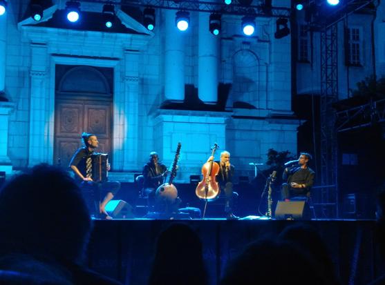 dans une lumière bleue sur une scène devant une porte d'église de style classique, 4 musiciens avec leurs instruments (accordéon, kora, violoncelle et saxophone soprano)