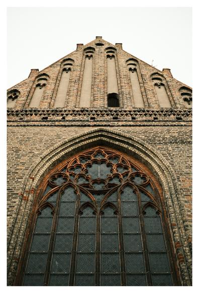 Blick nach oben auf die Fassade einer gotischen Backsteinkirche. Der spitze Giebel ist mit langen Spitzbogenblenden und kleinen Rundbogenöffnungen verziert. Darunter befindet sich ein großes Spitzbogenfenster mit feinem Maßwerk aus Backstein, in dem dunkel gerahmte Bleiglasscheiben eingesetzt sind.