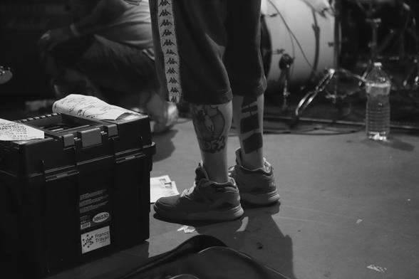 Photo en noir et blanc prise le 27 juin 2025 à Grrrnd Zero lors du premier soir de la troisième édition du festival Lixiviat : on y voit les pieds et les jambes du saxophoniste (!) du groupe Mental Hygiene Terrorism Orchestra et on remarque surtout un tatouage NoFX sur son mollet, ce qui quand on y pense est assez incongru dans le cadre d'un festival de musique consacré au grindcore et au powerviolence.