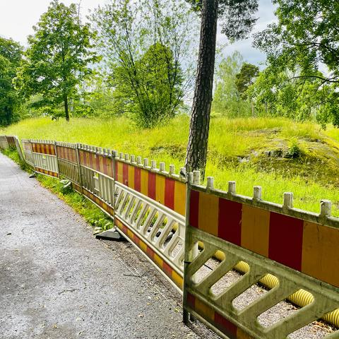 A street bordered by winding yellow and red safety barriers, with lush greenery and trees of a park in the background.