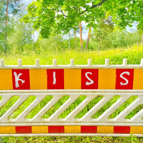 A fence with bright yellow and red safety stripe pattern and the word "KISS" painted in white on it, set against a lush, green backdrop of trees and grass.