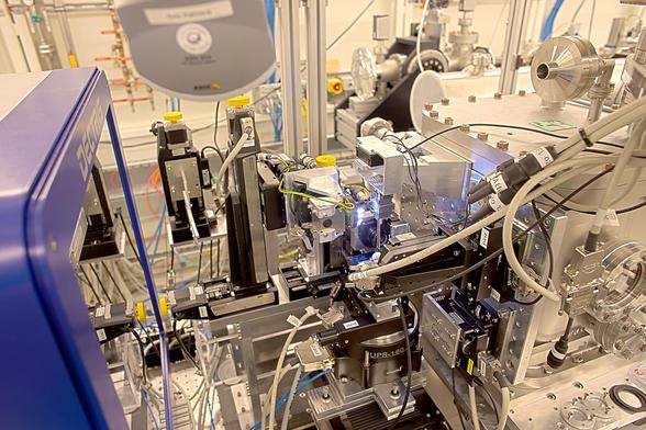 Foto, Blick ins Innere eines 3D-Röntgenmikroskopes. Es sind zahlreiche Motoren und Alu-Flächen zu sehen. Links ein großer Röntgendetektor, in dessen Schutzfolie sich Teile des Experimentieraufbaus wiederspiegeln; von oben ragt eine Webcam ins Bild; im Hintergrund Zuleitungen (Kabel, Luft, Gase) an der Wand. Viele Kabel, viel Gedöns, viel Flexibilität für die Forschung.