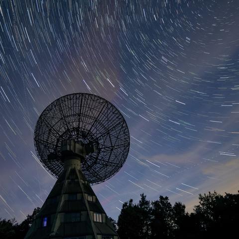A large satellite dish silhouetted against a starry sky with streaks of light, indicating star trails. The background features clouds and trees, creating a serene nighttime landscape.