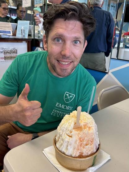 A photo of me with a bowl of Hawaiian shaved ice. The shaved ice is easily four times as tall as the bowl. I have my thumb up and am smiling.