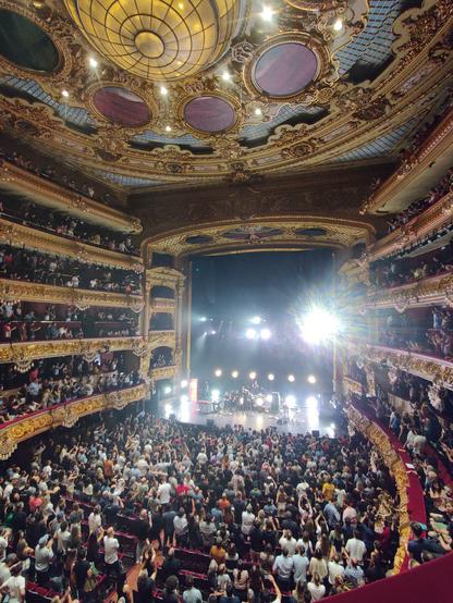An eclectic, baroque concert hall. View from one of the balconies towards the stage. A band can be seen on the stage. The whole hall is filled with spectators standing looking at the stage.
The concert hall is the Liceu in Barcelona, on stage is Andrés Calamaro and his supporting band.