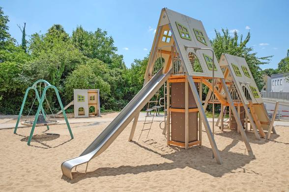 Spielplatz mit Klettergerüst in Form von Häusern, Rutsche und Spielhäuschen, umgeben von Bäumen.