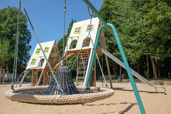 Ein Spielplatz mit einem großen Klettergerüst in Form von Häusern, Kletterleiter, Rutsche und schalenförmiger Nestschaukel auf Sand.