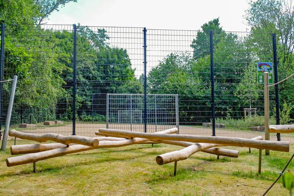 Holzspielplatz mit Balancierbalken aus Holzstämmen vor einem beschränkten Bereich mit Tor. Umgeben von grünen Bäumen.