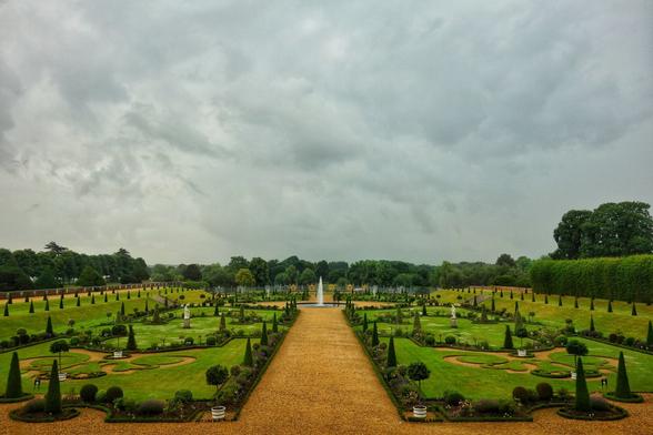 A wide shot captures the formal Privy Garden at Hampton Court Palace under a dramatic, cloudy sky. A long, straight, light-brown gravel path extends from the foreground into the distance, leading towards a tall, white water fountain at its end. Symmetrical green lawns, intricately shaped flowerbeds with low-trimmed hedges, and various topiaries flank both sides of the path. Scattered white statues are visible within the garden beds. In the background, a dense line of dark green trees forms a natural border, with glimpses of lighter green foliage beyond. The overall impression is one of an expansive, meticulously maintained Baroque landscape.