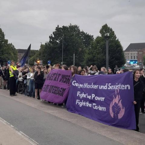 Blick auf die Demo von der Seite. Zwei Transpis sind zu sehen. Auf dem einen steht Gegen Macker und Sexisten fight the power fight the cistem