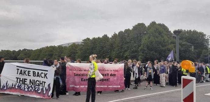 Demo von der Seite aus gesehen. Vorne wird eine Banner gehalten auf dem take back the night steht. An der Seite ist ein Banner auf dem Ob zur Kieler Woche oder im Alltag, sexistische Gewalt den Kampf ansagen drauf steht.