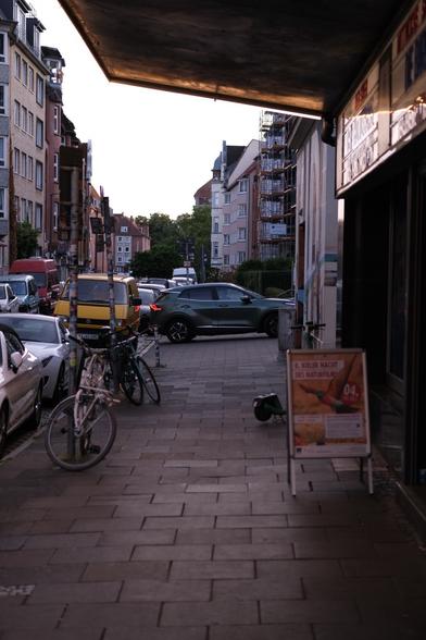Das Foto zeigt eine ruhige Straßenszene in einer Stadt neben dem Studio Kino in Kiel. Im Vordergrund ist ein Gehweg aus Steinplatten zu sehen, auf dem zwei Fahrräder an Straßenschlidern lehnen. Ein Schild mit der Aufschrift "8. KIELER NACHT DES NATÜRFILMS" steht am rechten Rand des Bildes. Dahinter steht ein Leihroller. Im Hintergrund sind mehrere Gebäude zu sehen, darunter Wohnhäuser mit Balkonen und ein Gebäude mit Gerüst. Parkplätze entlang der Straße sind mit verschiedenen Autos und einem Bulli belegt. Ein SUV steht in vollerbreite quer über dem Gehweg in einer Einfahrt. Die Straße ist kaum belebt und die Beleuchtung deutet auf Abendzeit hin.