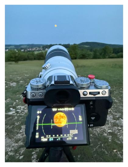 Foto im Hochformat. Ein Fotoapparat ist auf einem Stativ. Auf dem Display ist der Mond groß abgebildet. Am Fotoapparat ist ein langes, weißes Objekt, welches auf den Mond am Abendhimmel gerichtet ist. Im Vergleich zum Mond auf dem Display, ist er am Himmel nur ein kleiner Kreis.