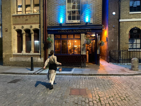Night shot Vanessa has a lantern and is seen from behind crossing the road. Many beautiful and historic windows in shot including the very inviting illuminated pub window
