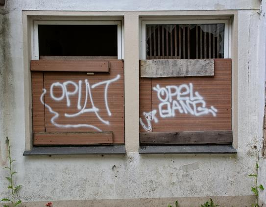 Two boarded-up windows in a ruin. One has ‘Opiat’ written on it, the other ‘Opel Gang’ in graffiti.