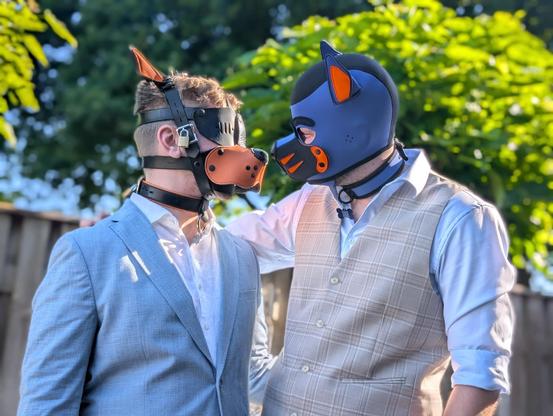 two men wearing their wedding suit and puppy mask. the left man has a light blue jacket with white shirt, and his mask is black and orange leather. the right man has a light brown checked vest over a light blue shirt, his mask is blue and black neoprene with orange accents. there are some trees in the background.