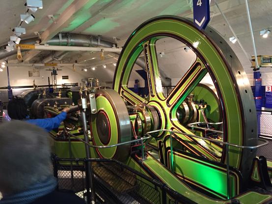 Eine industrielle Dampfmaschine mit einem großen grünen Schwungrad ist in einem Museum ausgestellt. Eine Person, die ein blaues Hemd trägt, interagiert mit den Maschinen, während Besucher aus der Ferne beobachten. Die Umgebung verfügt über ein industrielles Dekor und informative Schilder.