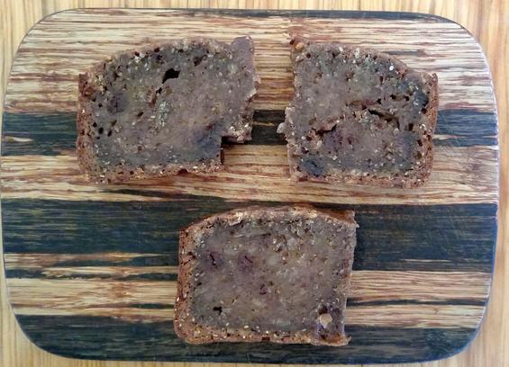 photo of a wooden plate with three small pieces of banana bread on it. They are dense, dark and kinda wet. They have little bits in them of a darker colour (blueberry snacks cut up into small bits). Two of the pieces used to be one but broke apart in the middle, where the hole from the kneading hook of the breadmaker was.