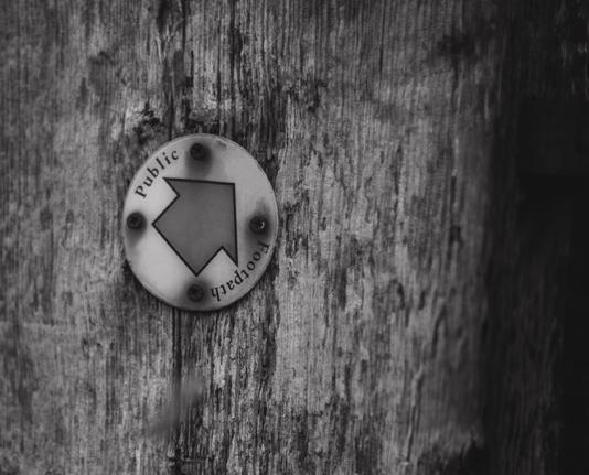 A black and white photograph of a wooden post with a round sign with the words Public Footpath on it.
