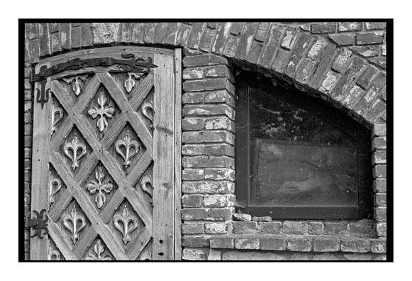A close-up of an ornate wooden door with carved fleur-de-lis patterns, set in a brick wall next to an arched window. The textures of wood and weathered bricks are accentuated by the grain of Ilford Delta 3200 black and white film.