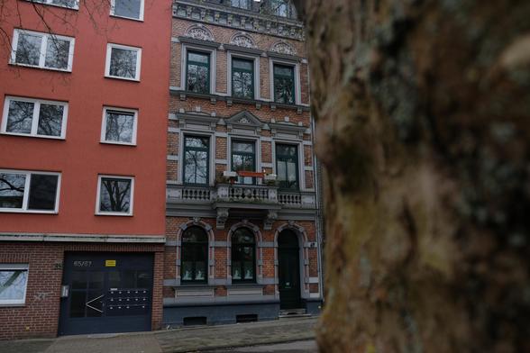 A row of houses. On the left, a house with a plastered red facade from the second floor upwards, with a brick wall underneath. To the right of the house is another one with a brick wall and an old mini balcony on the first floor. A blurred tree adjoins the edge of the house, which is in the foreground close to the camera.
