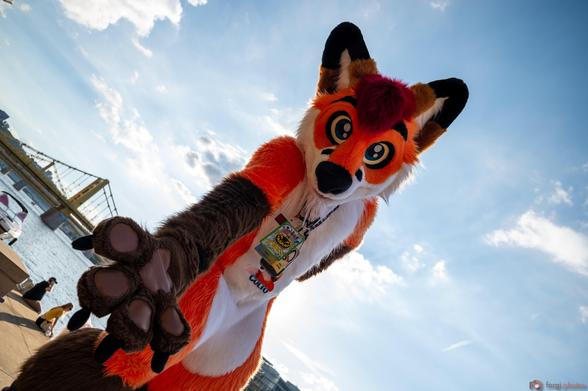 Colto Fox (orange, white, brown and red fox fursuiter) standing in full fursuit with one paw behind his back and the other reaching out in front. He's standing in front of a bridge in Pittsburgh, PA. He's wearing convention badges and the sky is clear with a few clouds.