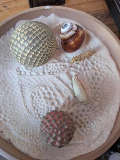 a small dish containing fine white sand, two textured balls that roll to make patterns in the sand, a sea shell, and two toy figures (a spotted leopard and a white seal)
on the counter at a cafe