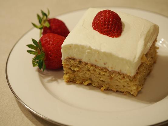 Ein Stück des Tres Leches Kuchens auf einem weißen Kuchenteller. Das Kuchenstück wurde mit einer halben Erdbeere garniert. Neben dem Kuchenstück liegen weitere Erdbeeren.