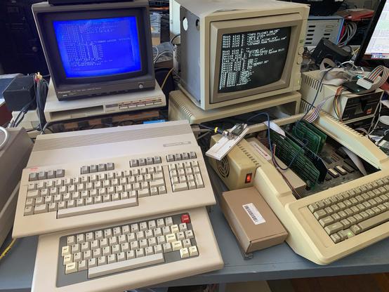 Commodore 128 in 64 mode sharing monitor with TRS-80 CoCo 3 with 128 stacked on top of CoCo. On the right is a IIe with an Apple Color 100 RHB monitor.